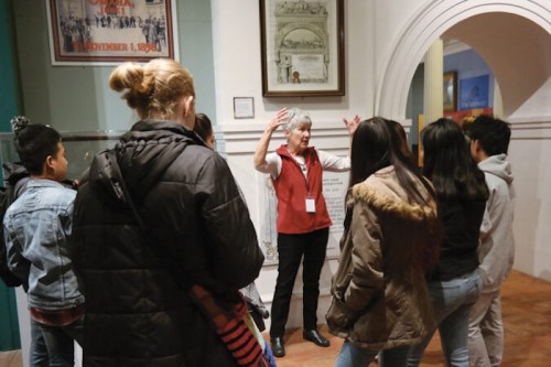 A museum guide explains an exhibit to a group of attentive visitors in a historical setting.