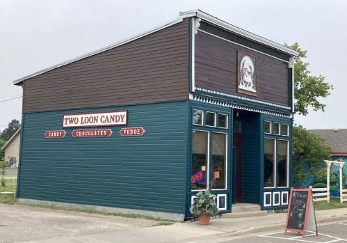 A colorful candy shop named "Two Loon Candy," featuring a sign and large windows, with a chalkboard outside.