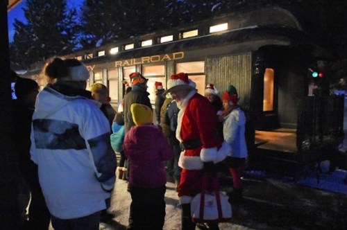 A group of people, including Santa, gather outside a vintage train car decorated for the holidays in a snowy setting.