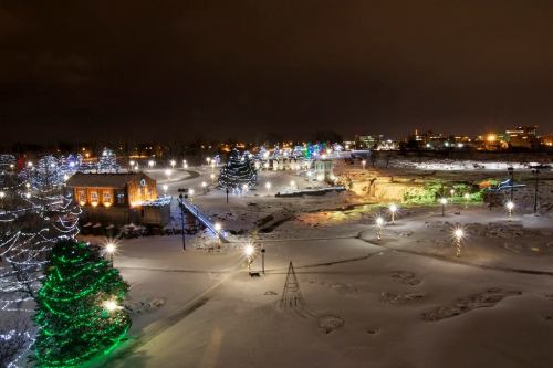 A snowy landscape at night, featuring illuminated trees and buildings, creating a festive winter atmosphere.