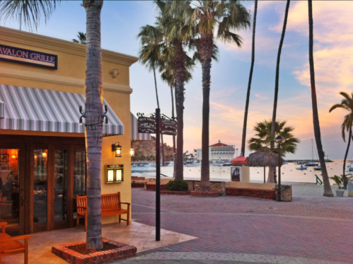 A coastal scene featuring a restaurant, palm trees, and a sunset over the water with boats in the background.