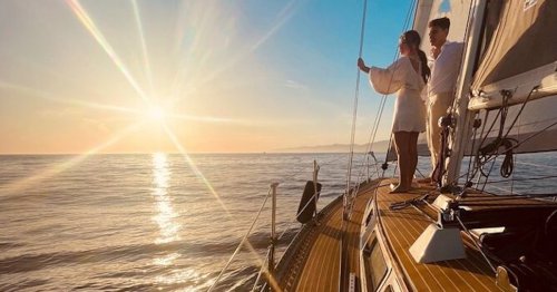 A couple stands on a sailboat, gazing at a sunset over calm waters, with sunlight reflecting on the ocean.