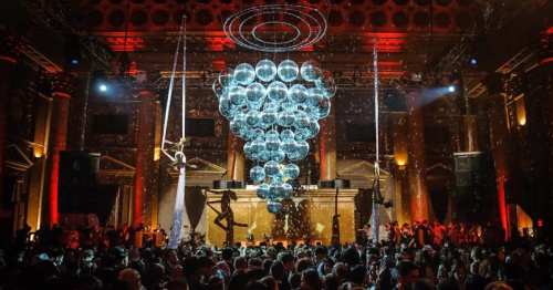 A vibrant party scene with a large chandelier of disco balls, aerial performers, and a lively crowd in a grand venue.