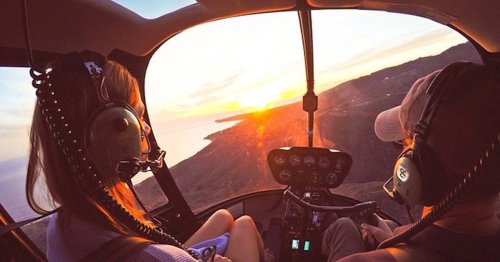 Aerial view from a helicopter cockpit at sunset, with two passengers wearing headsets enjoying the scenery.