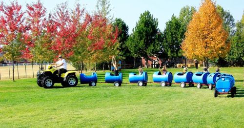 A yellow ATV pulls a colorful train of blue barrels on wheels through a grassy field with autumn trees in the background.