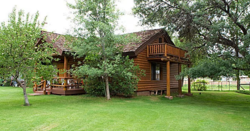 A charming log cabin surrounded by greenery and trees, featuring a balcony and a well-maintained lawn.