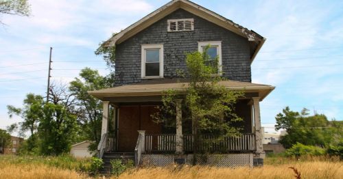 Abandoned two-story house with overgrown vegetation and a weathered exterior, set against a blue sky.