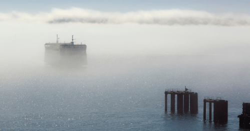A large ship emerges from fog, with distant piers partially visible in the calm water.