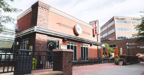 A retro-style diner with a clock on the exterior, surrounded by outdoor seating and modern buildings in the background.