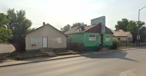 Two houses side by side on a street; one is light gray and the other is green with a sign. Trees are visible nearby.