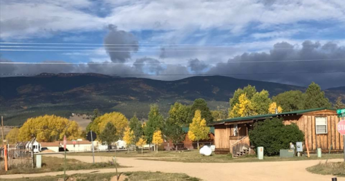 A scenic view of a rural area with mountains, colorful trees, and a wooden building under a cloudy sky.