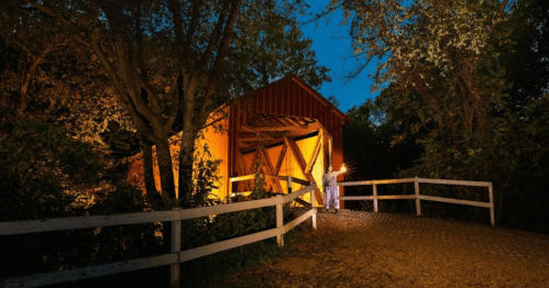 A lit-up covered bridge at night, surrounded by trees and a white fence, with a person holding a lantern nearby.
