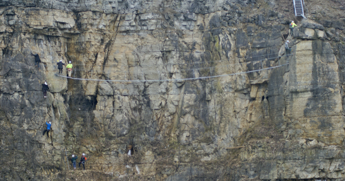 A group of climbers traverses a rocky cliff using a rope bridge, with a ladder visible in the background.