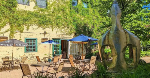Outdoor patio with tables and umbrellas, surrounded by greenery and a large sculpture in the foreground.