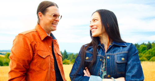 Two smiling individuals in denim jackets stand in a field, enjoying a lighthearted moment together.