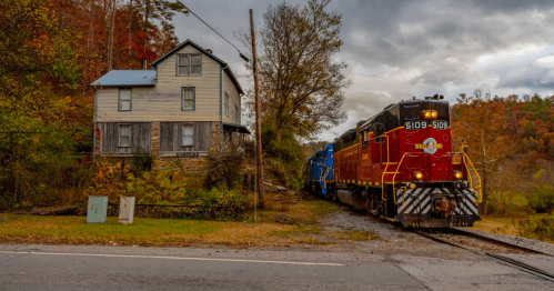 A red and black train passes by a weathered house surrounded by autumn foliage and a cloudy sky.
