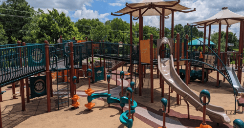 A colorful playground with slides, climbing structures, and shaded areas surrounded by trees and blue skies.