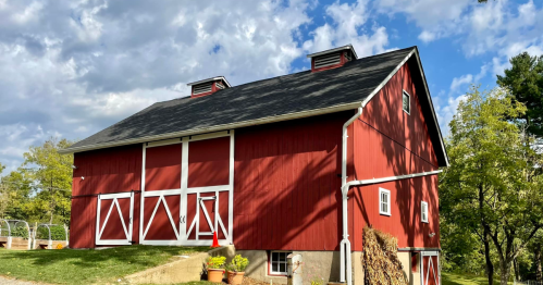 A red barn with white doors and a gray roof, surrounded by trees and a cloudy blue sky.