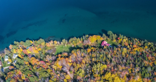 Aerial view of a colorful autumn landscape with a lake and a red-roofed house surrounded by trees.