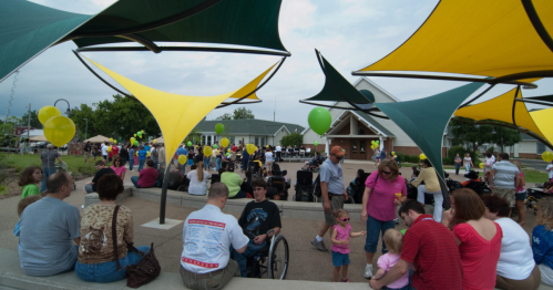 A community event with colorful canopies, people socializing, and balloons in a park setting.
