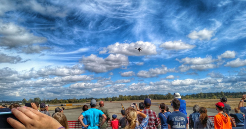 A crowd watches an aircraft perform in the sky, with fluffy clouds and blue skies in the background.