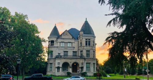 A grand, historic mansion with turrets, surrounded by trees, against a sunset sky.