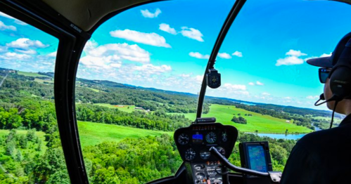 A pilot's view from a helicopter cockpit, flying over lush green fields and a blue sky with scattered clouds.