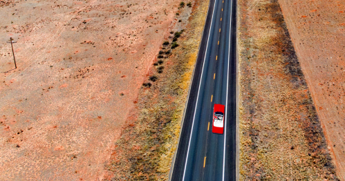 Aerial view of a red car driving on a long, empty road through a barren landscape.