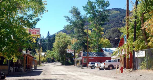 A quiet dirt road lined with trees and buildings in a small town, with mountains in the background.