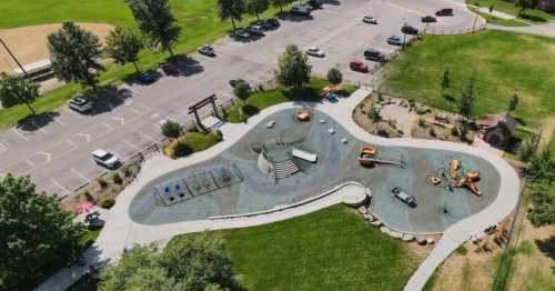 Aerial view of a playground with various equipment, surrounded by green grass and a parking lot in the background.