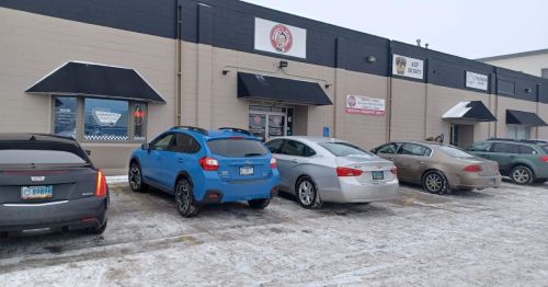 A row of parked cars in front of a commercial building with signage, on a snowy day.