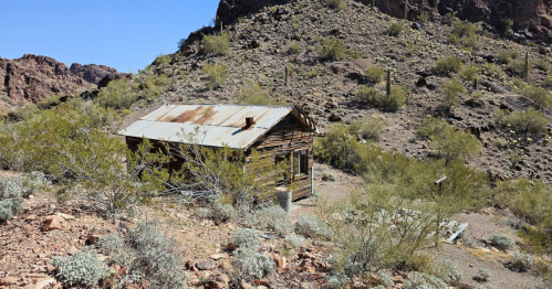 Abandoned wooden cabin in a desert landscape, surrounded by sparse vegetation and rocky hills under a clear blue sky.