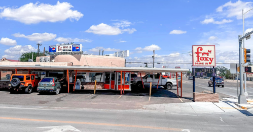 A drive-in restaurant with a red and white sign, parked cars, and a clear blue sky in the background.