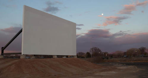 A blank movie screen stands on a dirt mound under a colorful sunset with a visible moon in the sky.