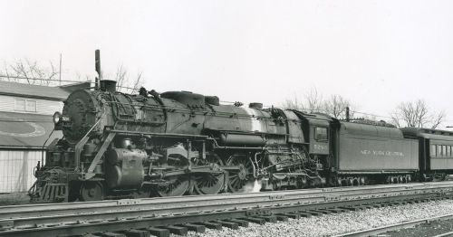 A vintage steam locomotive with a passenger car, parked on tracks near a building, in black and white.