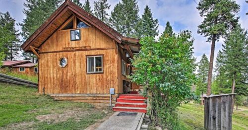 A rustic wooden cabin with red steps, surrounded by tall trees and greenery under a cloudy sky.