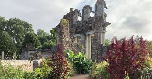 Ruins of a stone structure surrounded by lush greenery and vibrant plants under a cloudy sky.