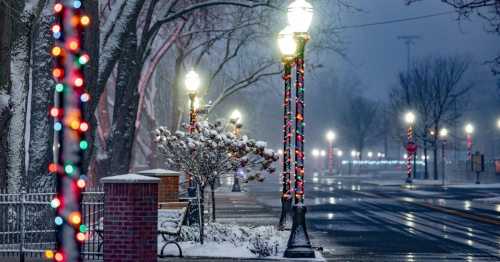 A snowy street scene at night, with colorful holiday lights on lampposts and trees lining the road.