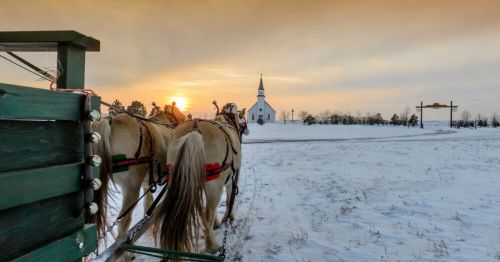 Horses in harness stand in the snow, with a church and sunset in the background.