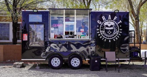 A black food truck with "Craft Donuts & Coffee" signage, serving customers in a sunny outdoor setting.