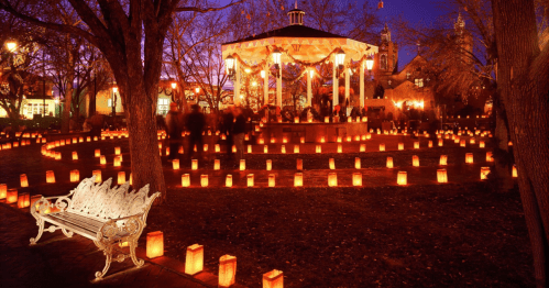 A festive park scene at dusk, featuring a gazebo surrounded by glowing lanterns and a white bench in the foreground.