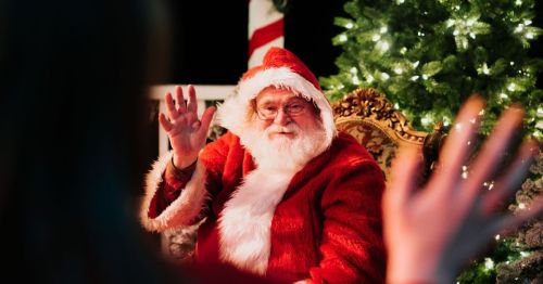 Santa Claus waves from his chair, surrounded by festive decorations and a Christmas tree.