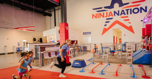 Children and an instructor run through an obstacle course at a vibrant indoor ninja training facility.