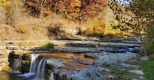 A serene waterfall cascades over rocky ledges, surrounded by autumn foliage and a calm river.
