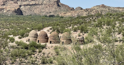 Four stone kilns nestled in a desert landscape, surrounded by green vegetation and mountains in the background.
