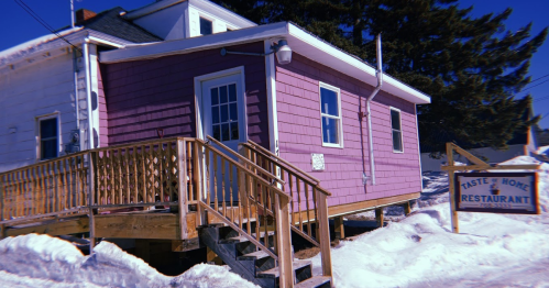 A purple restaurant building with a wooden ramp, surrounded by snow, featuring a sign that reads "Taste of Home."