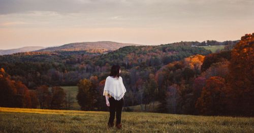 A person stands in a field, gazing at a colorful autumn landscape with rolling hills and trees.