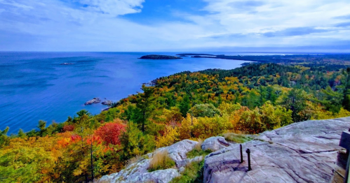 A scenic view of a coastline with vibrant autumn foliage and a calm blue lake under a partly cloudy sky.