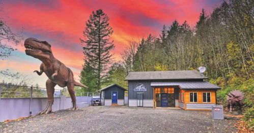 A dinosaur statue stands near a modern building against a vibrant sunset sky, surrounded by trees.
