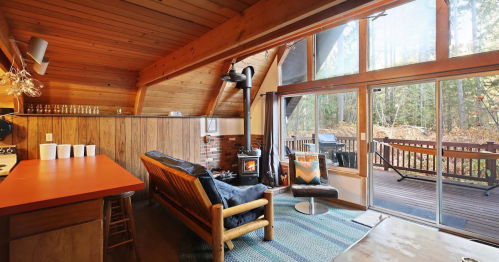 Cozy wooden cabin interior with a sofa, stove, and large windows overlooking a deck and forested area.